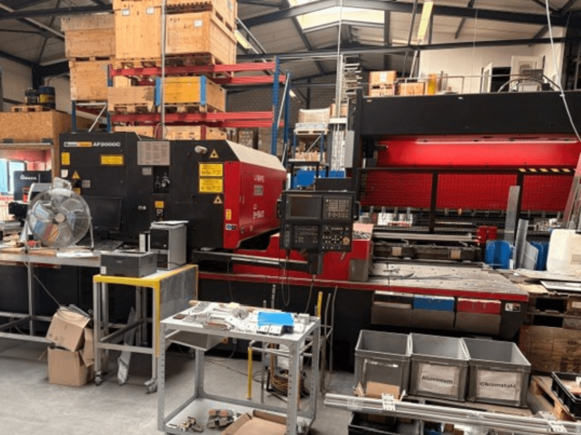 AMADA LC-2415αIII CO₂-laserlõikemasin töökoda-keskkonnas, millel on näha esivaade, juhtpaneel ja tööala.