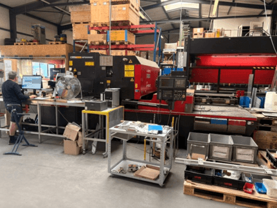 AMADA LC-2415αIII CO₂-laserlõikepink töökoda-keskkonnas, kus esiplaanil on näha arvutitöökoht.