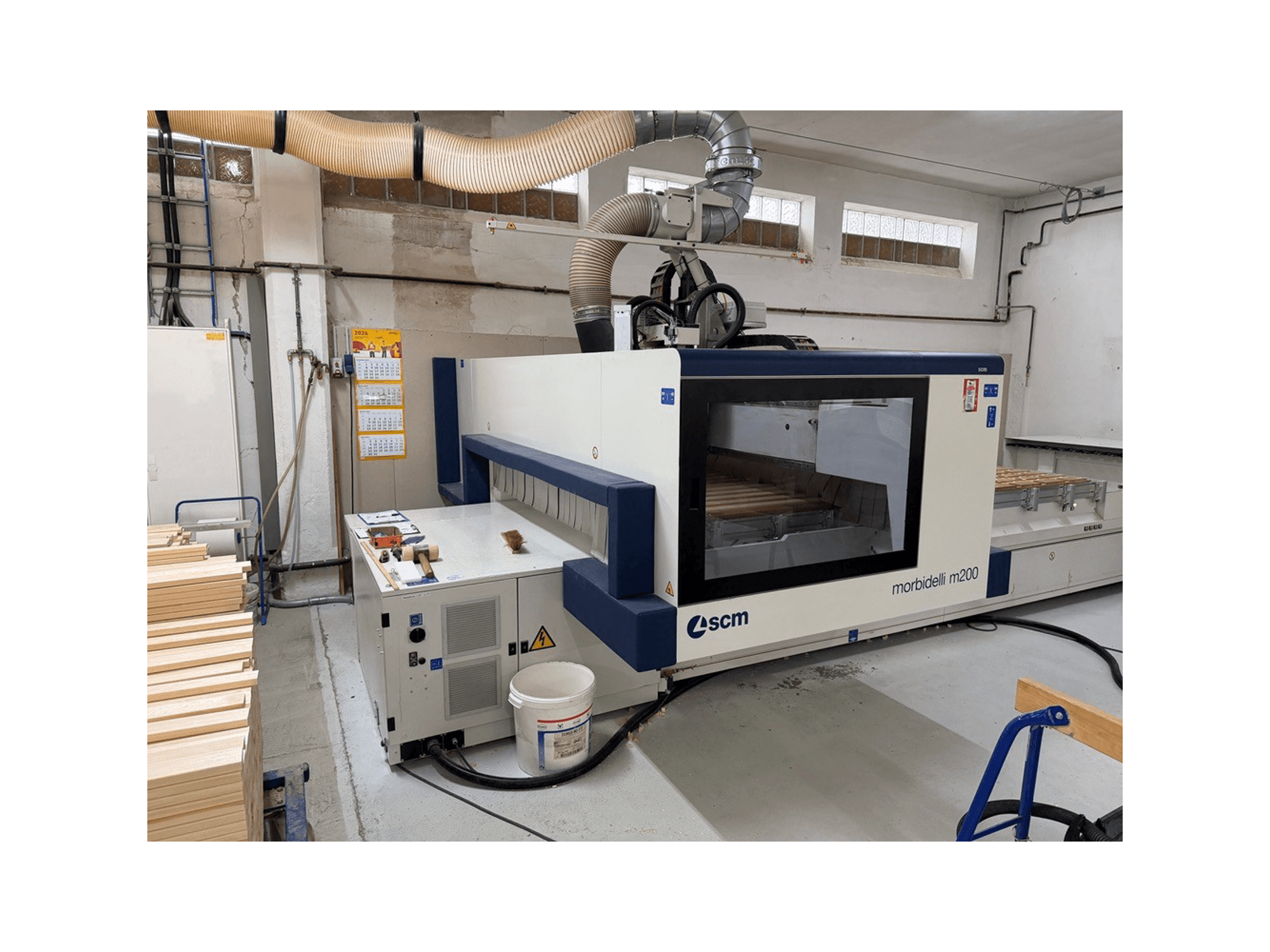 Töökojas asuv SCM Morbidelli M200F CNC-töötlemiskeskus, külgvaade, kus laual on puitdetailid ja kohal asub ventilatsioonisüsteem.