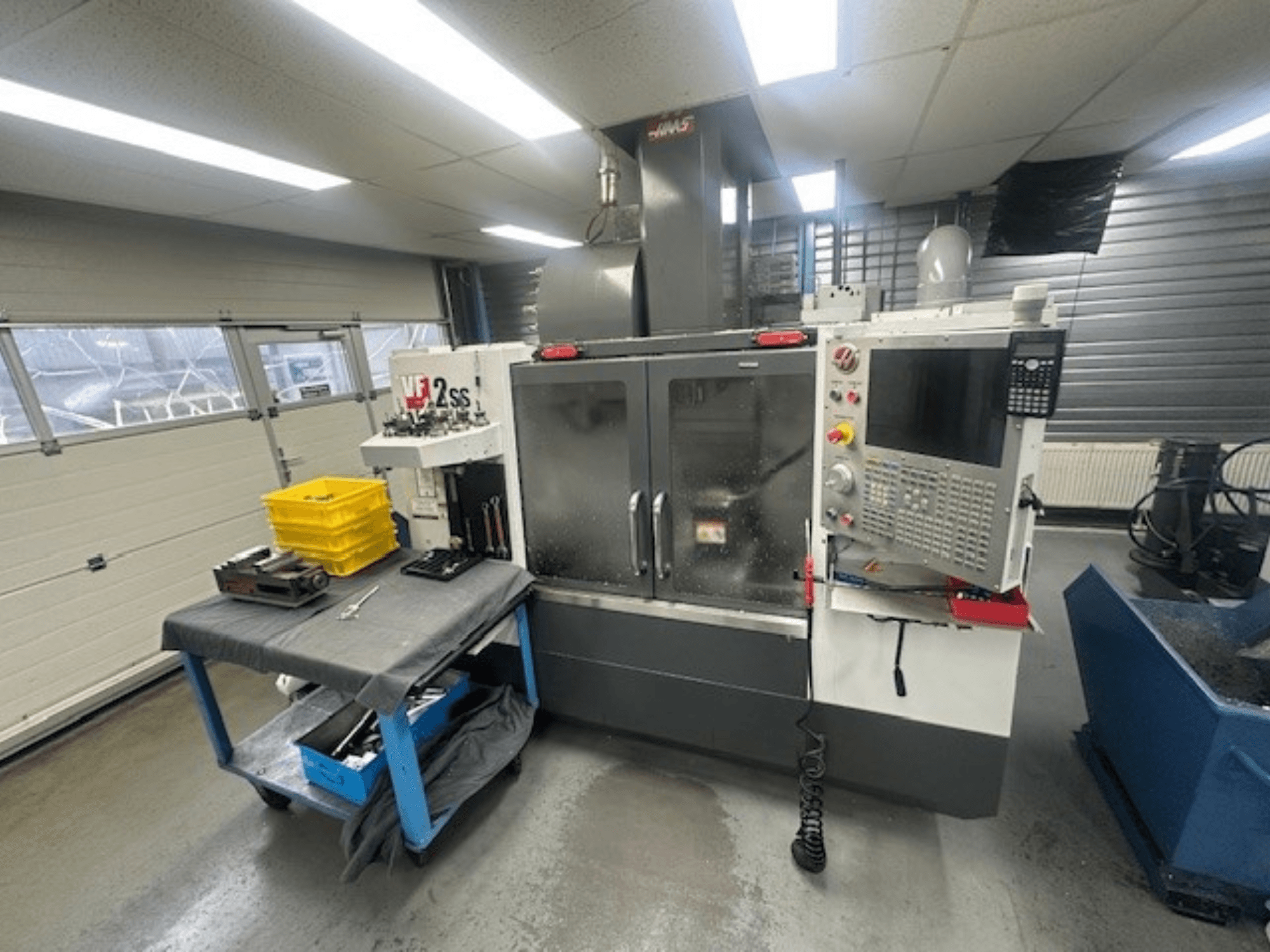 HAAS VF-2SS vertikaalse töötluskeskuse esiosa koos juhtpaneeli, töölaua ja tööriistade hoiukohta sisaldava paigutusega.