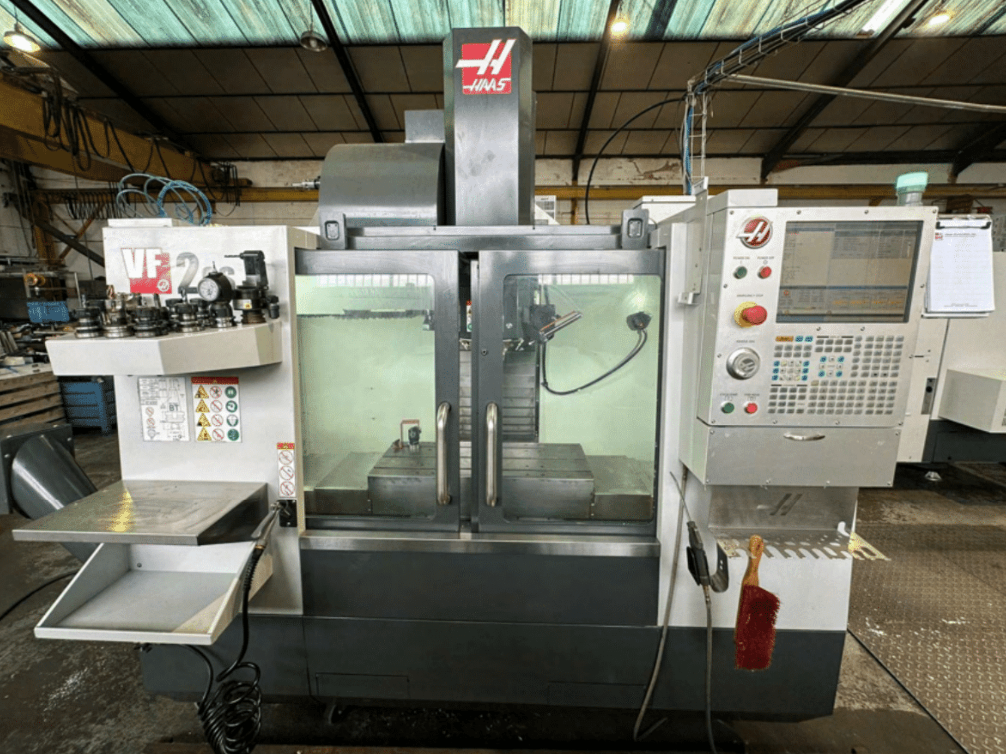 Haas VF2 CNC-pingi esivaade, kus on näidatud juhtpaneel, tööriistahoidjad ja tööruum koos ohutussiltide ja pintsliga.