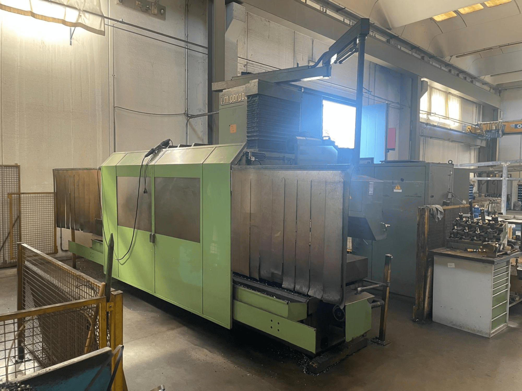 Roheline tööstuslik masin nähtavate metallosadega, esiplaanil; juhtpaneel, ohutushoiatused ja töökoht lähedal.