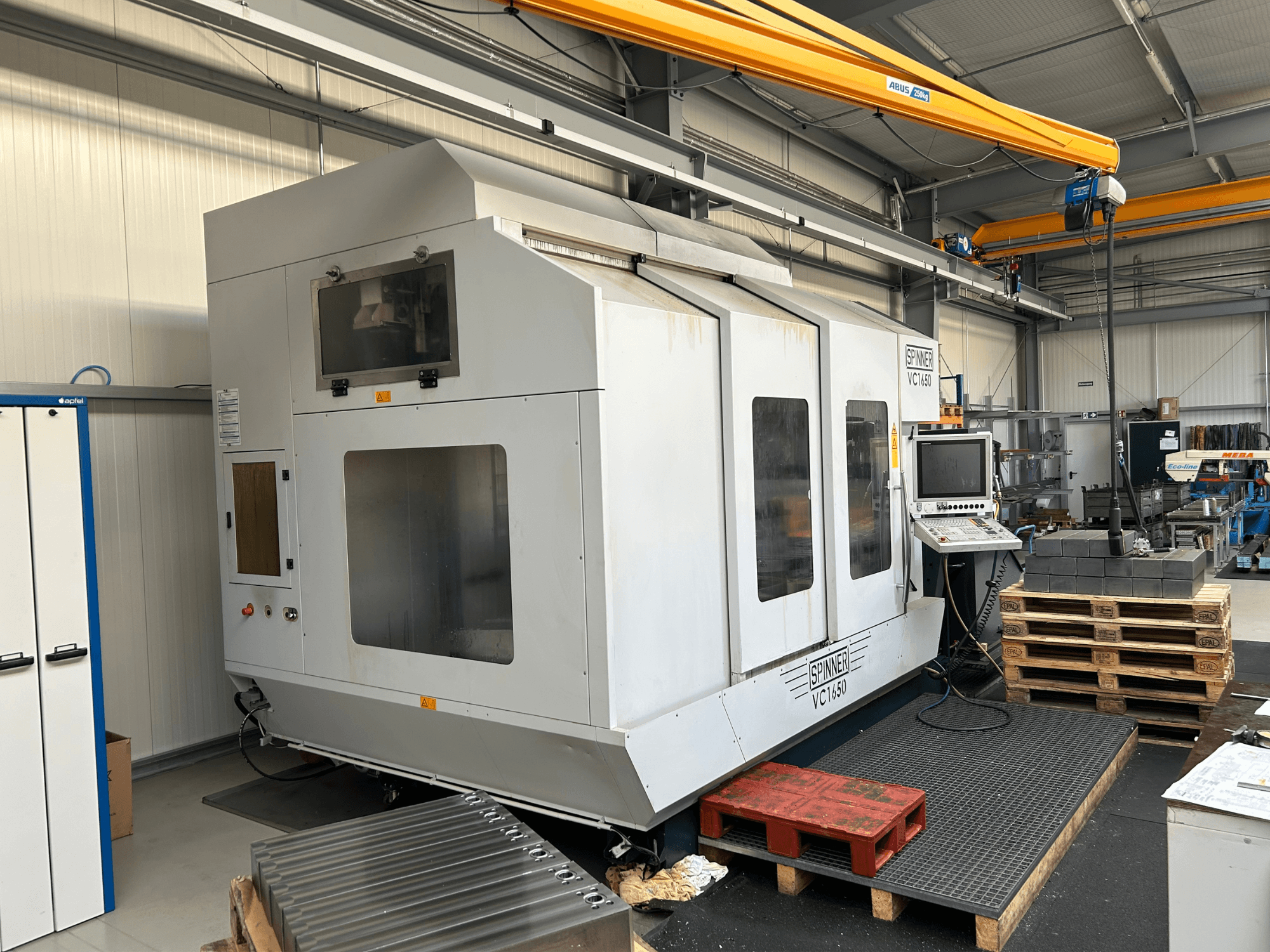 CNC-töötlusmasin Spinner VC 1650, esivaade, juhtpaneel, suured aknad ja läheduses virnastatud kaubaalused töökojas.