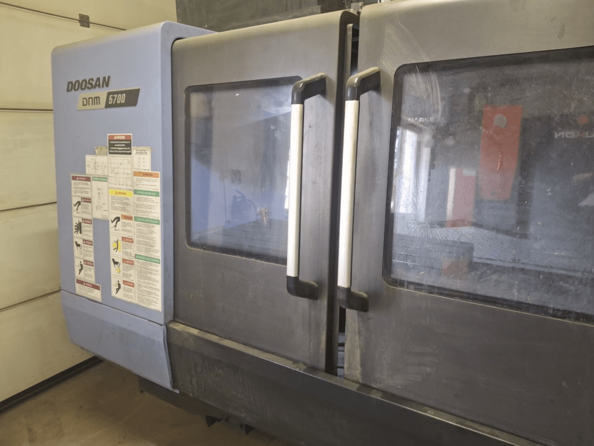 DOOSAN DNM 5700 vertikaaltöötluskeskuse esiplaan, kus on näha kaks ust ja juhtpaneelid koos tööjuhistega.