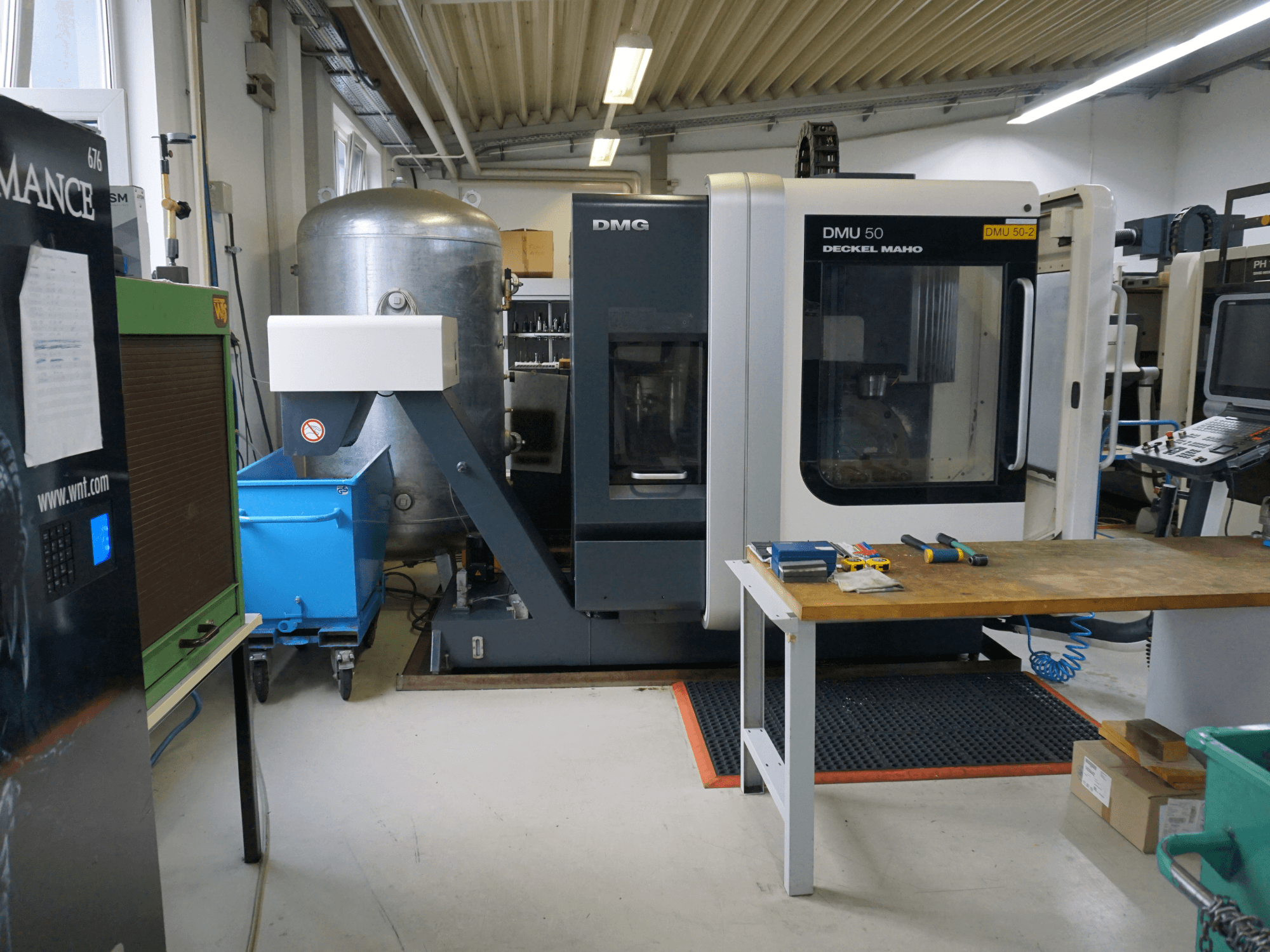 DMG DMU 50 freespink töökojas, esiplaanil, tööriistad puidust laual ja sinine ratastega prügikast lähedal.