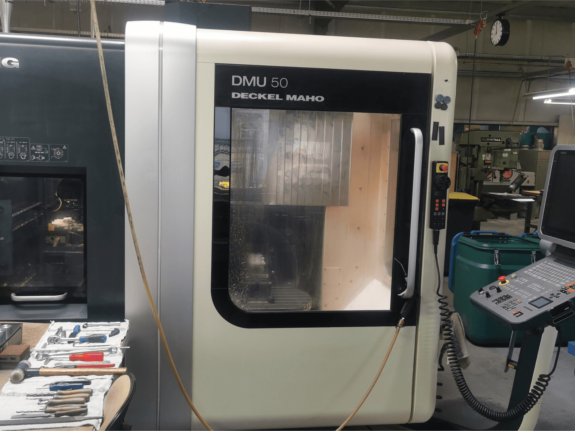 DMU 50 deckel maho CNC-tööpink ettepoole vaadatuna, kus on näha juhtpaneel ja läbipaistev uks. Tööriistad on lähedal.