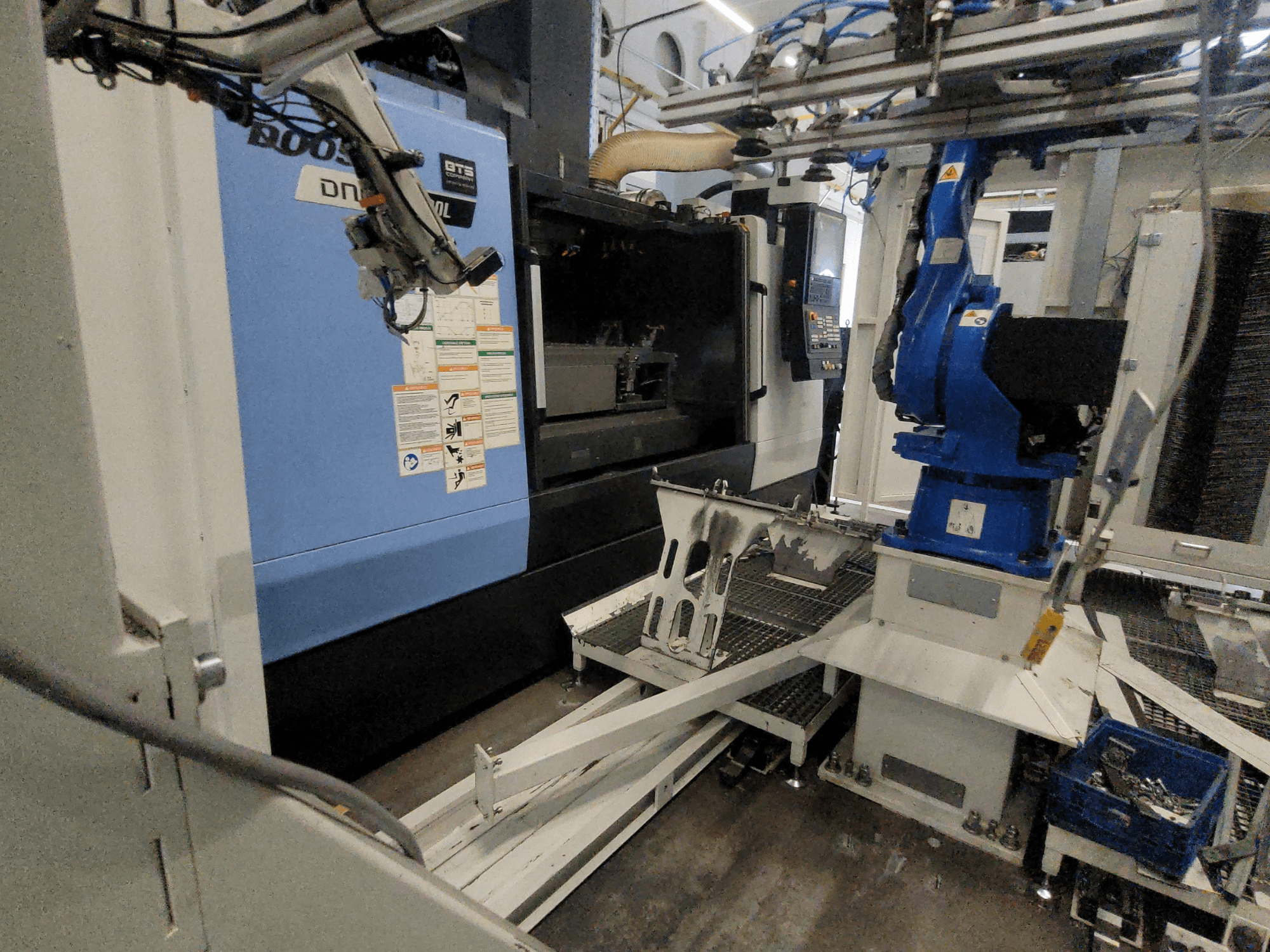 DOOSAN DNM 6700L vertikaalse töötlemiskeskuse külgvaade, millel on näha robotkäsi ja juhtpaneel ning mis illustreerib masina seadistust.