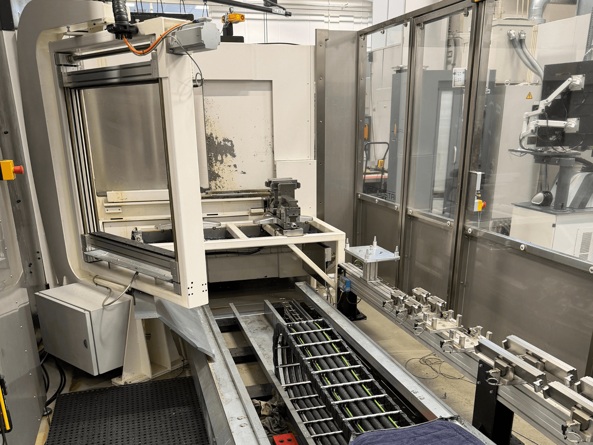 Automaatne tööstuslik tööjaam, kus on näha robotkäsi, rööbastee süsteem ja juhtpaneel külgvaates.