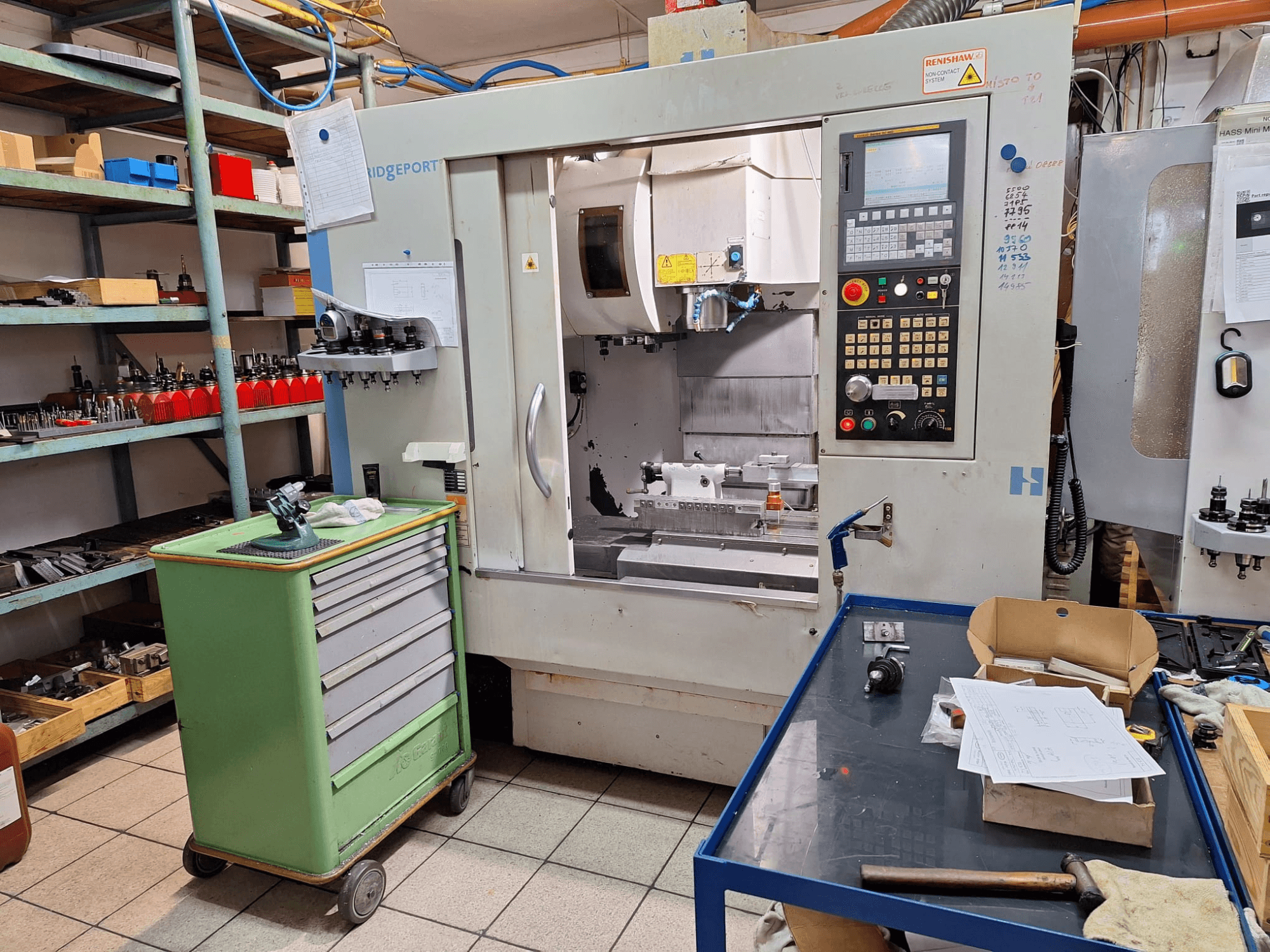 Hardinge Bridgeport GX 710 vertikaalse töötlemiskeskuse esivaade koos juhtpaneeli, tööriistade ja riiulitega töökoda keskkonnas.
