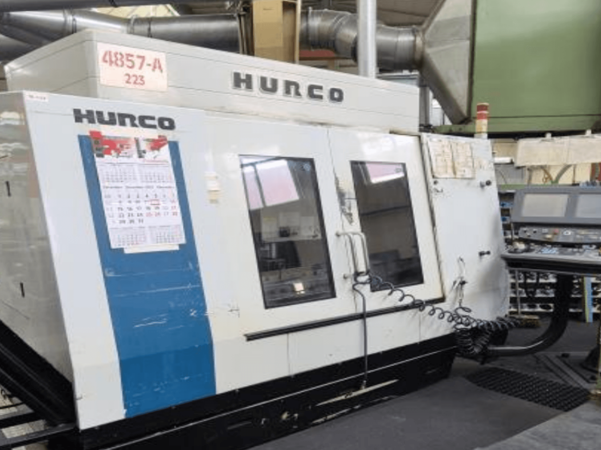 Hurco VMX 42 vertikaalne töötluskeskus esiplaanil, millel on juhtpaneel, kalender ja metallist välisdetailid.