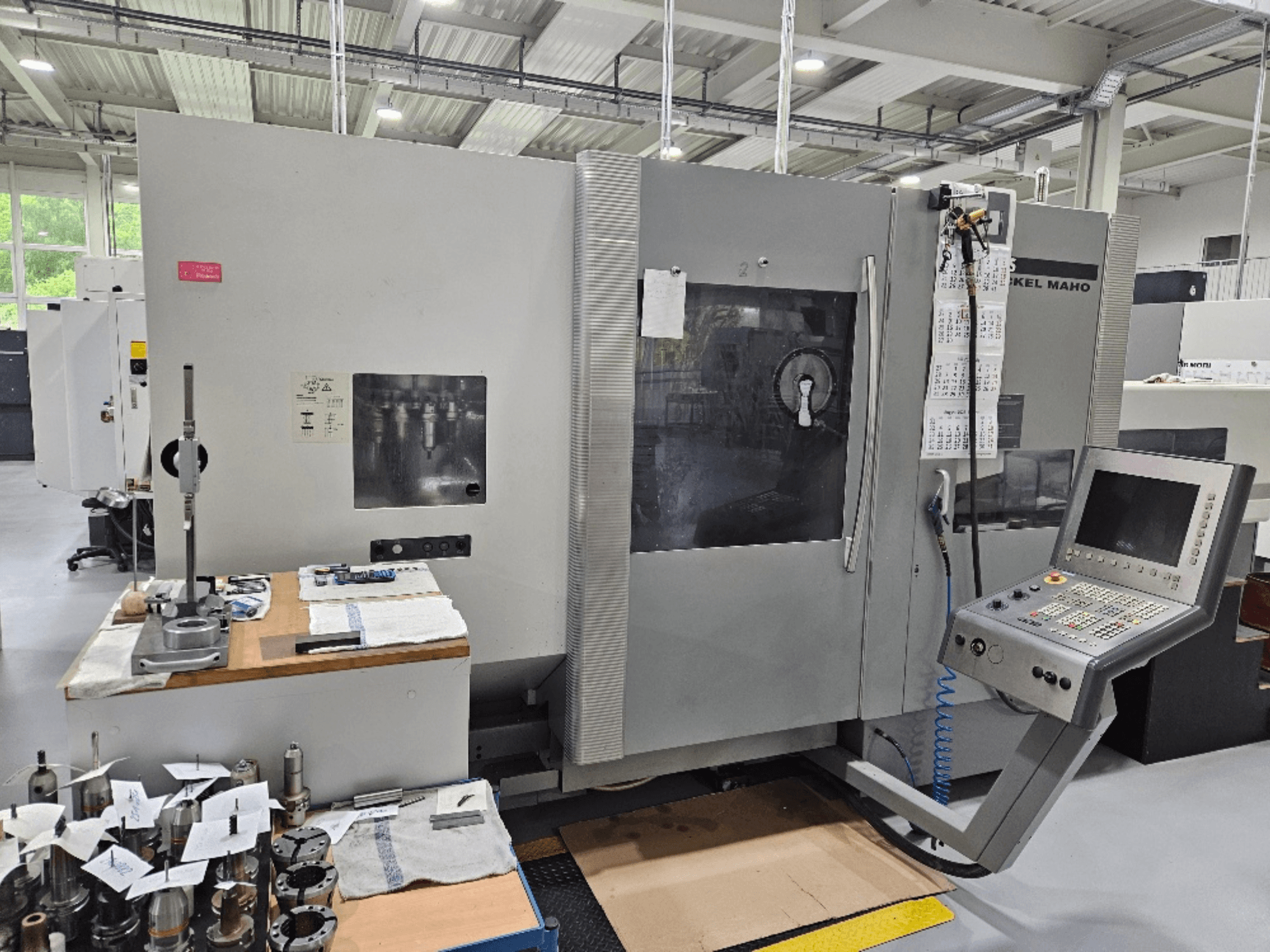 DMG Mori DMU 70 masina esivaade juhtpaneeliga, tööriistad puidust laual ja erinevad töödeldavad detailid lähedal.