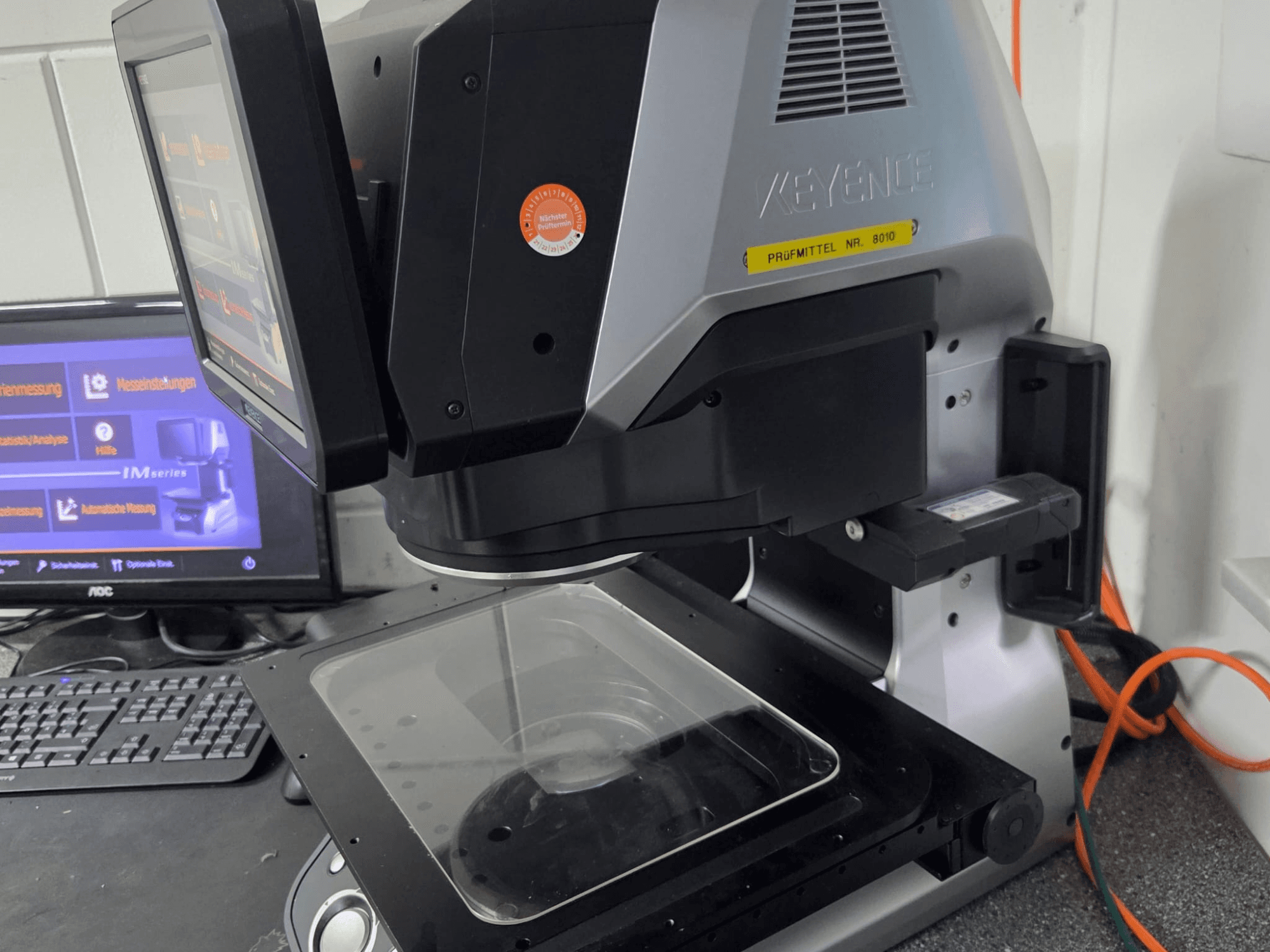 KEYENCE IM 7030 optiline mõõtmismasin, esiplaanil koos puutetundliku ekraani ja klaasist mõõtmisplaadiga.