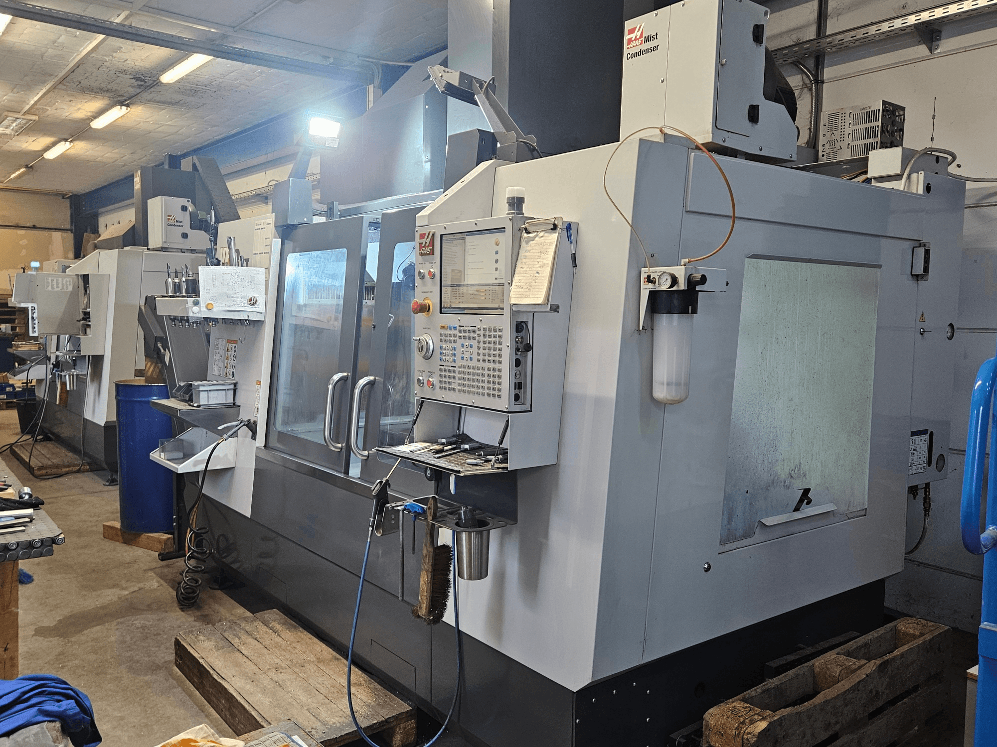 Haas VF-3YT/50 vertikaalne töötlemiskeskus töökodas, vaade eest, millel on näha juhtpaneel, tööriistahoidikud ja udu kondensaator.