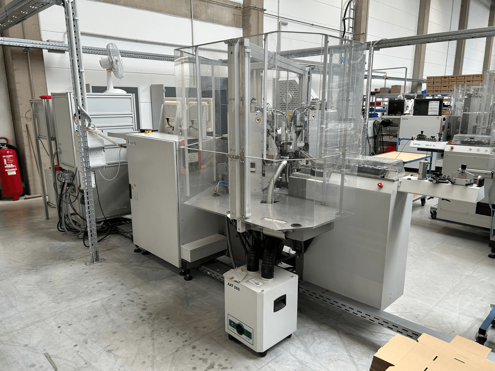 KOMAX Alpha 433 L automatiseerimisseadmed tehase keskkonnas. Seadmel on läbipaistev ohutuskate ja mitmesugused juhtpaneelid.
