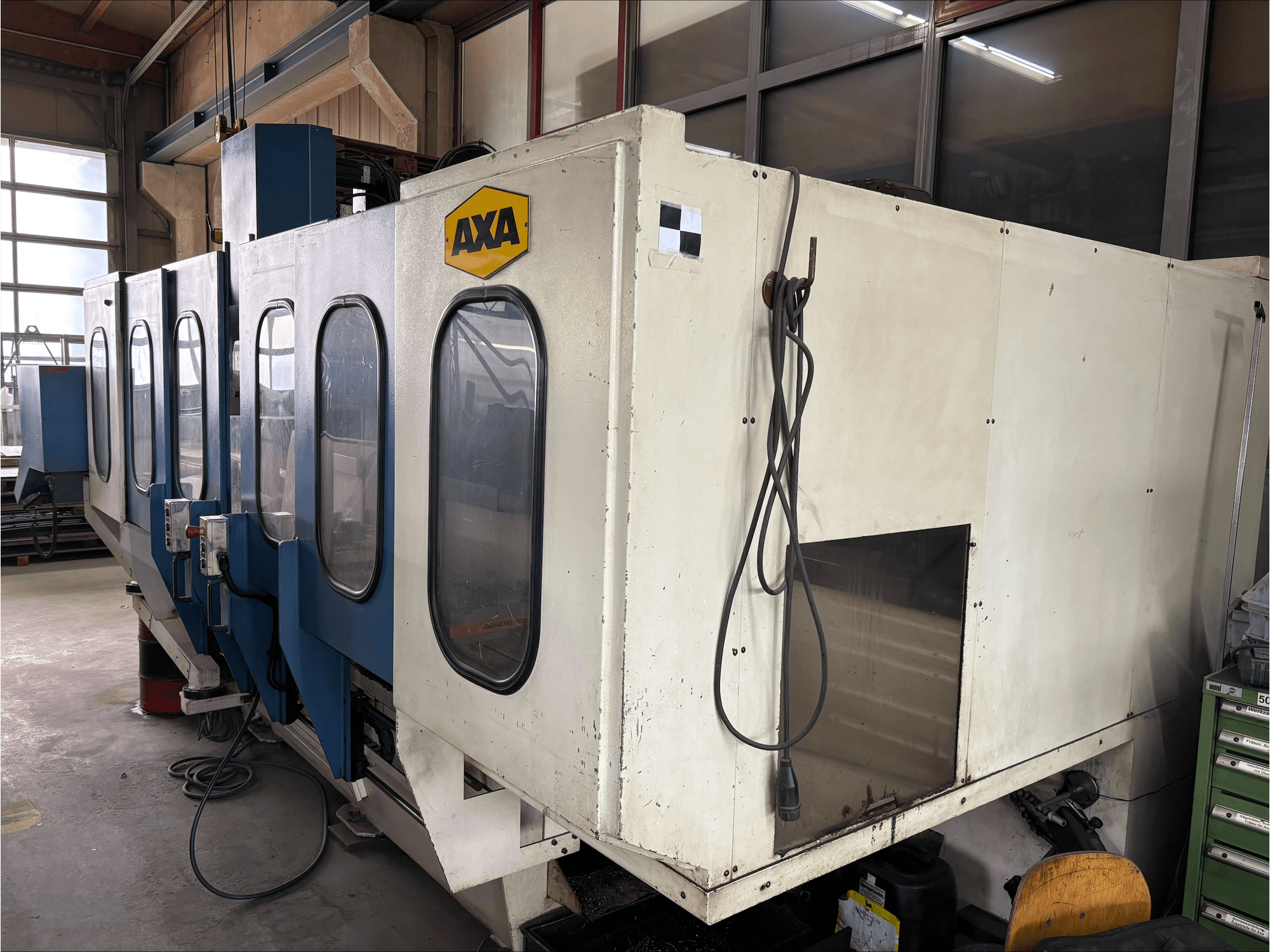 AXA VSC-1-M/C vertikaalne töötluskeskus, esiplaanil, millel on suur valge kapp ja sinine juhtpaneel.