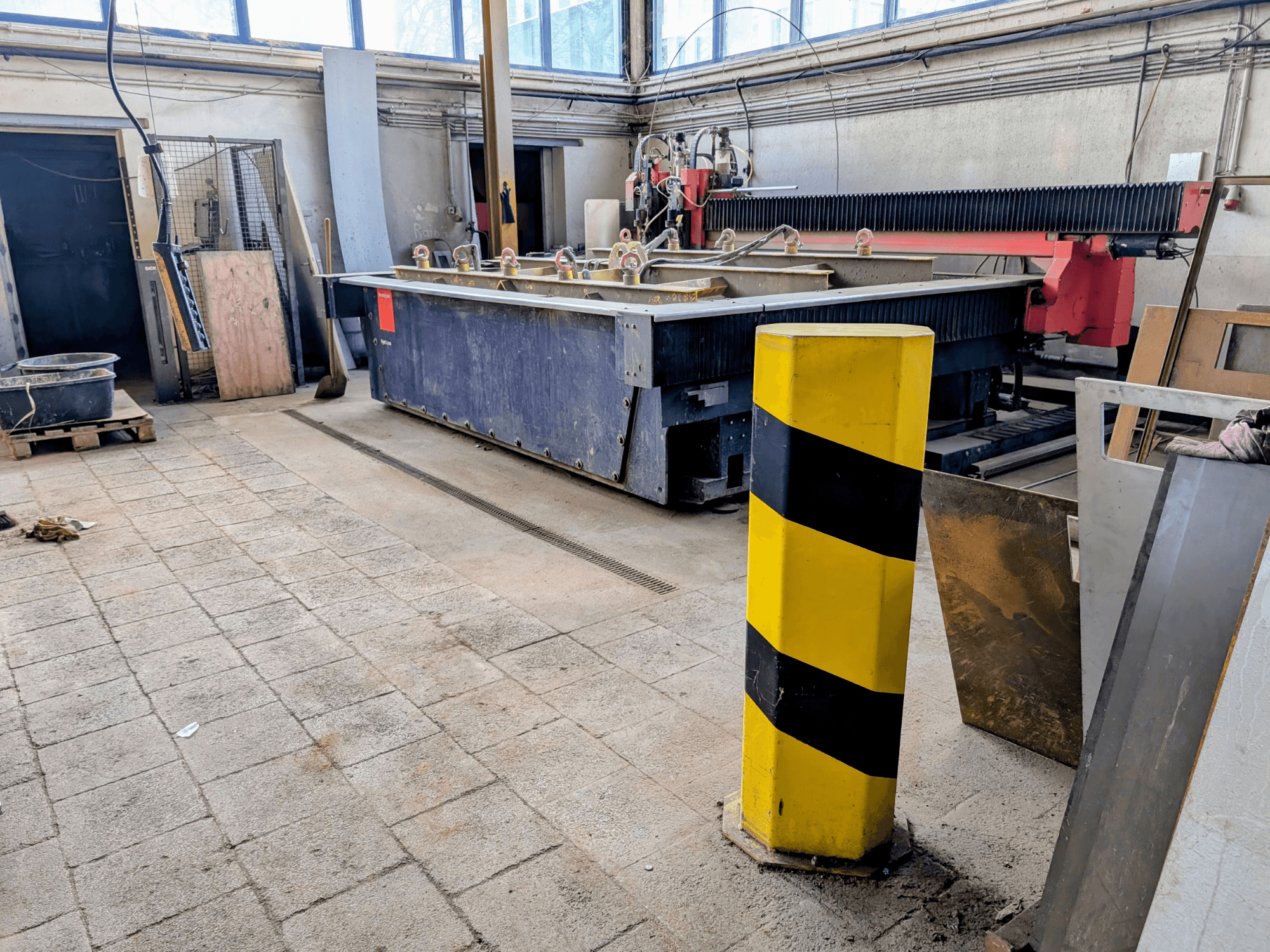 Bystronic Byjet L 2030 vesilõikepink töökojas, külgvaade, kollane ohutuspulk ja ümbritsevad materjalid.