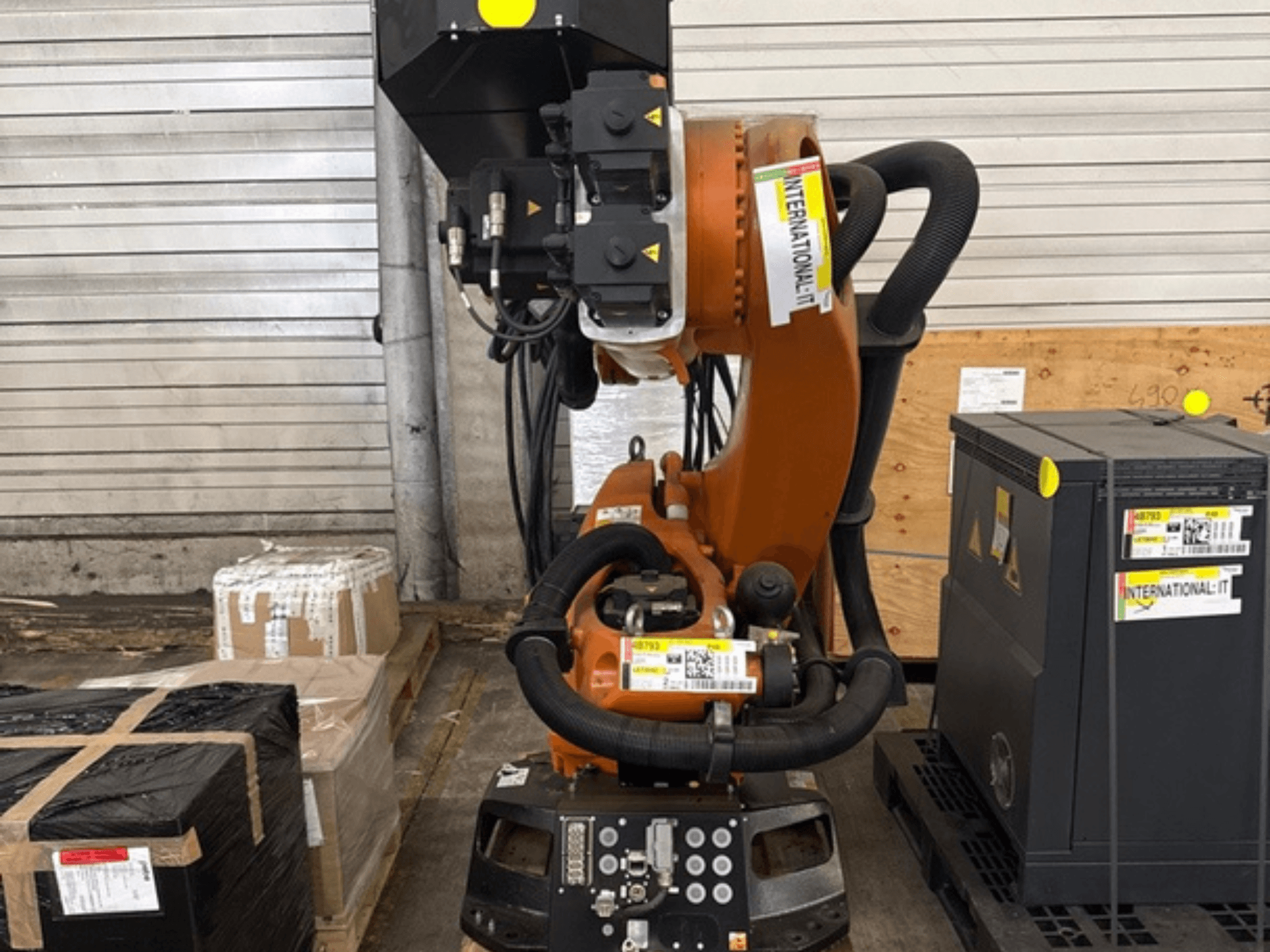 KUKA KR 210 R2700 lisarobotkäsi eestvaates, eksponeeritud kaubaalusel pakendite ja seadmete kõrval.