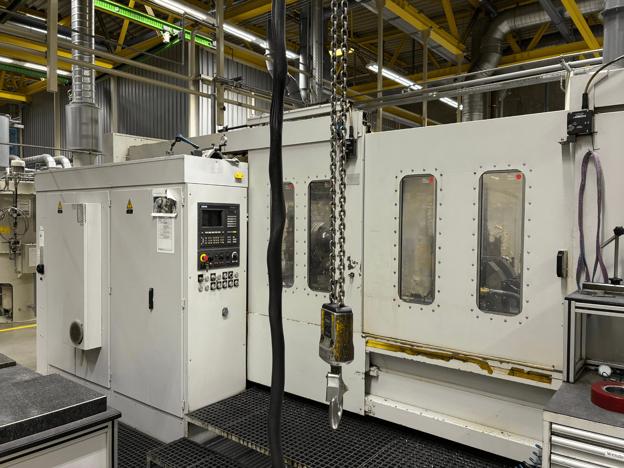 Tööstuslik CNC-masin koos juhtpaneeliga, külgvaade; aknad, ketid ja metallkomponendid tehases.